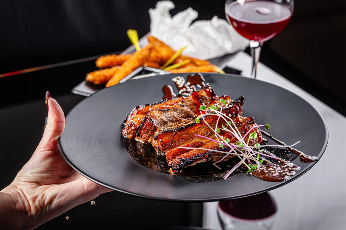 Glazed pork ribs served on a black plate with microgreens and wine