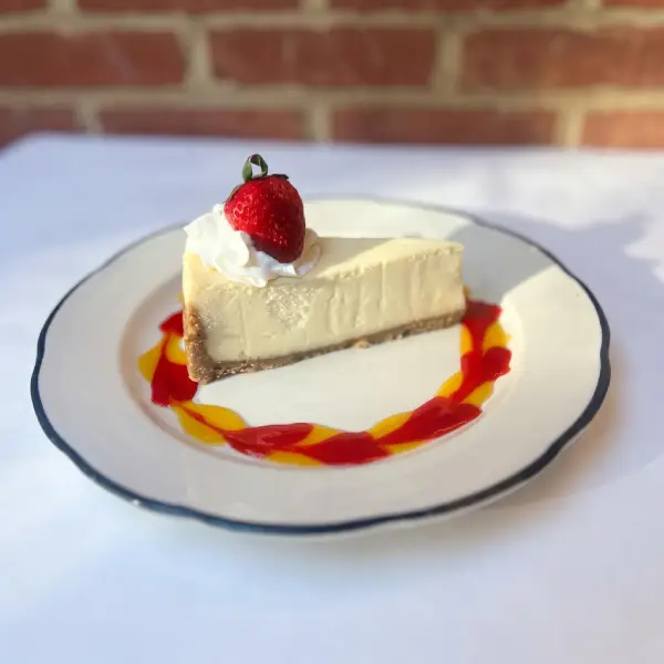 Slice of cheesecake topped with a strawberry, with raspberry and mango swirl sauce